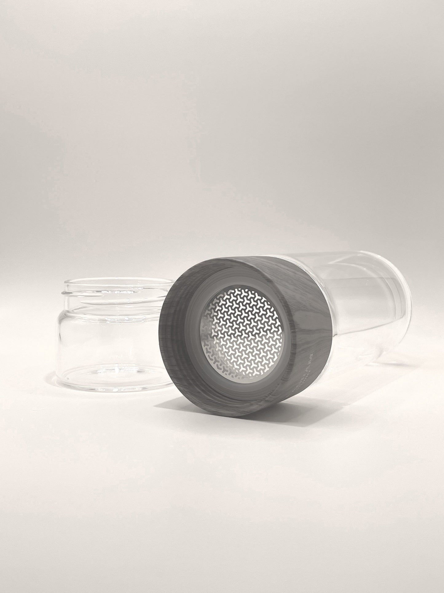 The Brew & Go Tea Bottle by My Store is a transparent, cylindrical double-wall tea bottle featuring a grey lid with a perforated metal mesh design. Displayed horizontally with the lid placed beside it, this bottle is perfect for tea drinkers who appreciate a seamless tea experience on the go. Its minimalistic white background enhances its elegance.