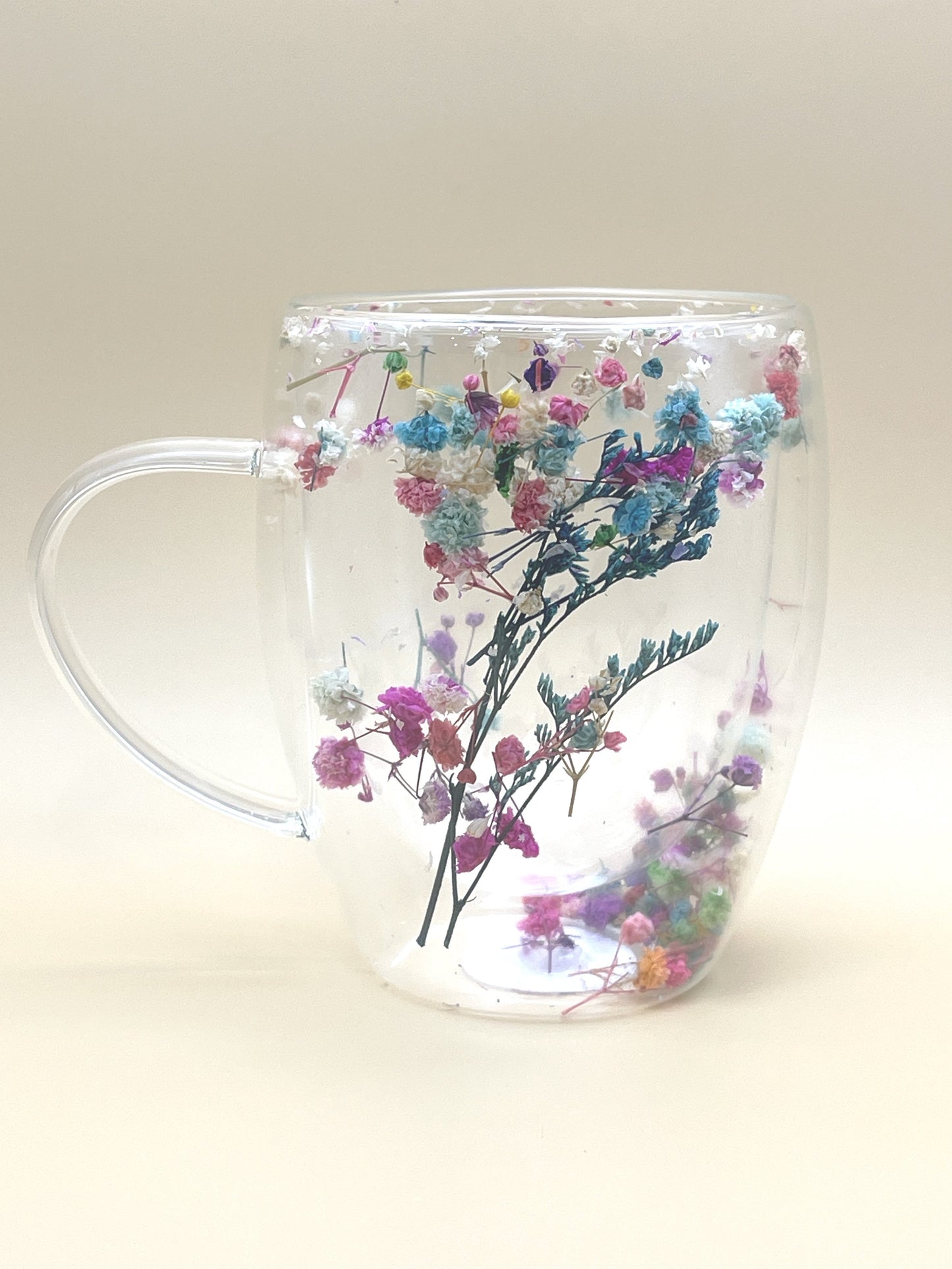 The BloomVista Floral Cup by My Store is a clear, double-walled flower cup filled with water, showcasing an enchanting arrangement of delicate multicolored dried flowers. Inside, pink, blue, white, and purple blossoms with small green stems are beautifully suspended, creating a vibrant and whimsical display against a plain background.