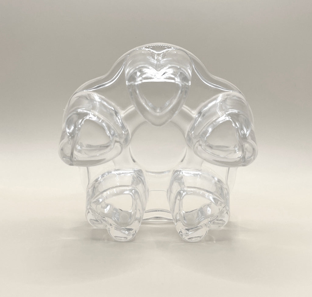A clear, heart-shaped plastic ring, featuring five heart-shaped loops arranged in a circular pattern around a central opening, is reminiscent of the charm of the CozyGlow Tea Warmer by My Store. The object is set against a plain, light background.