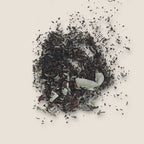 A scattered pile of Bear's Hug Tea from My Store on a light background. The mix includes small, dark tea leaves, dried flower petals, and white, thin slices of dried fruit or other botanical ingredients. Perfect for relaxation and promoting digestive health with its natural blend of antioxidants.