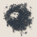 A circular arrangement of My Store's English Breakfast loose black tea leaves is displayed on a beige background. The tea leaves, rich in antioxidants for heart health, vary in size and form a textured, uneven pattern. Some smaller fragments are scattered around the main cluster.