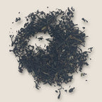 A circular arrangement of My Store's English Breakfast loose black tea leaves is displayed on a beige background. The tea leaves, rich in antioxidants for heart health, vary in size and form a textured, uneven pattern. Some smaller fragments are scattered around the main cluster.