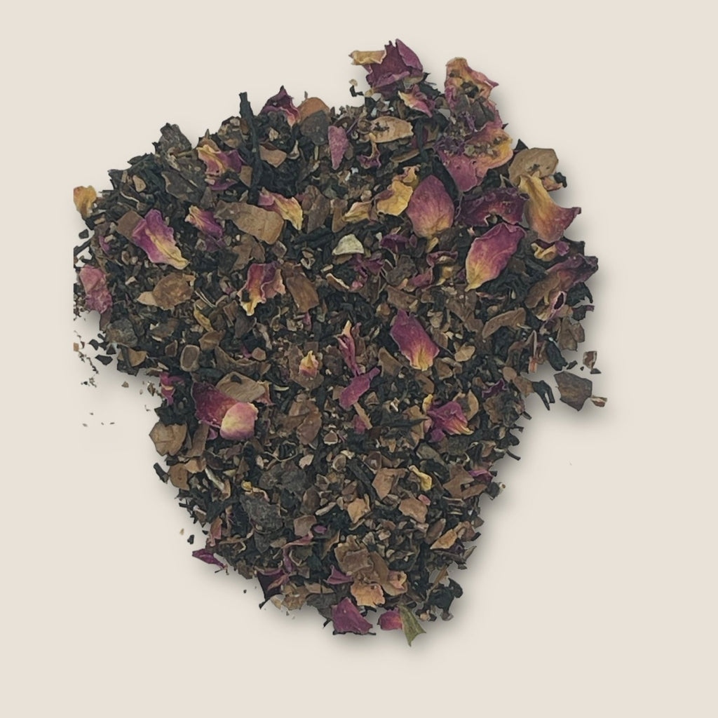 Cocoa Tea by My Store features loose tea leaves mixed with dried rose petals, scattered on a plain, light background. The dark cocoa tea leaves and the pink rose petals create a contrasting and visually appealing blend, rich in antioxidants that support overall well-being.