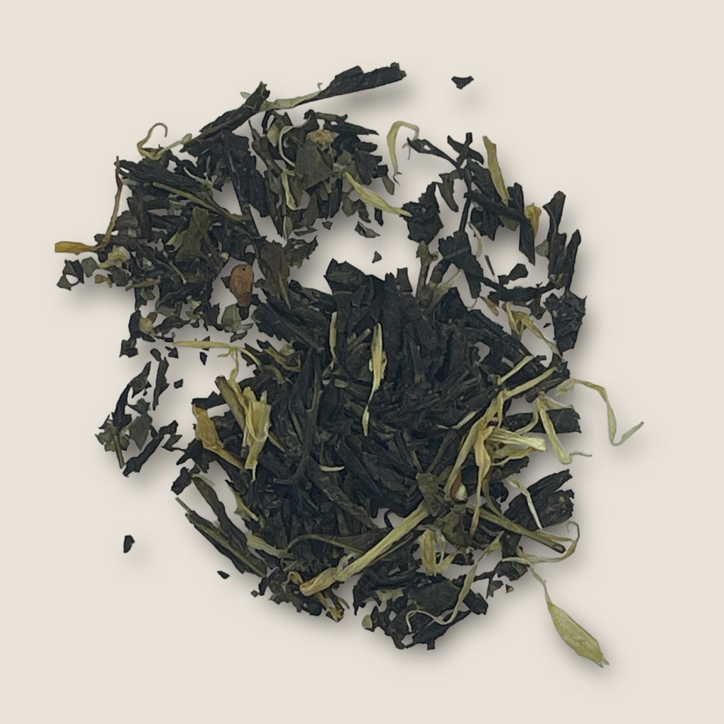 Apple Mojito, a loose leaf green tea from My Store known for its antioxidants and immune support, is blended with dried leaves and flower petals on a light beige background. The tea leaves are dark green and slightly curled, interspersed with light green and yellowish petals, creating a contrast in color and texture.