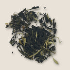 Apple Mojito, a loose leaf green tea from My Store known for its antioxidants and immune support, is blended with dried leaves and flower petals on a light beige background. The tea leaves are dark green and slightly curled, interspersed with light green and yellowish petals, creating a contrast in color and texture.