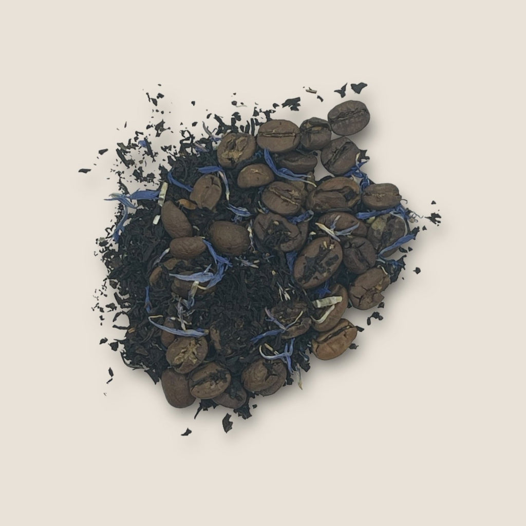 A visually textured blend of Espresso Martini by My Store features dark black tea leaves mixed with roasted coffee beans and small purple petals on a light beige background. The combination offers a striking contrast of deep colors and vibrant purple, promising an energy boost with each sip.