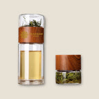 The image features two glass tea infusers from My Store. The taller Brew & Go Tea Bottle has a double-wall design with a wooden cap and is filled with tea and water. The smaller container, also capped with wood, holds loose tea leaves. Perfect for the tea drinker on the go, the background is light beige.