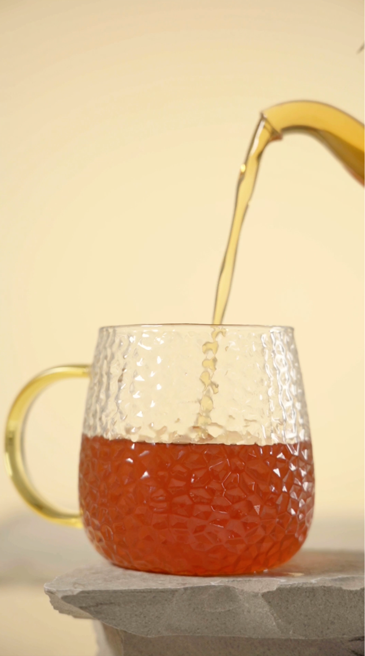 A clear textured glass mug with a gold-toned handle and spout, partially filled with amber tea, with more tea being poured into it from a matching gold-toned vessel against a warm cream background.