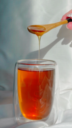 A clear double-walled glass cup filled with amber honey, with a wooden spoon holding a dripping stream of honey above it. A hand with white nail art is visible holding the spoon on a white draped fabric background.