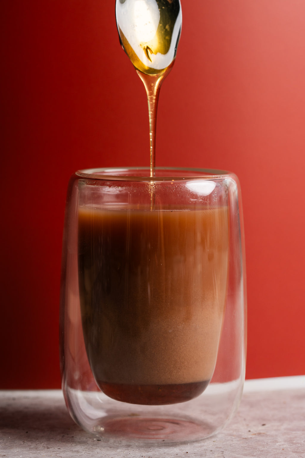 A double-walled clear glass cup filled with a brown liquid, with a spoon above pouring a glossy golden syrup or honey into the center. The background is a solid red studio backdrop.