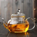 A transparent Crystal Clarity Teapot from My Store, made of high borosilicate glass and holding 600ml of amber-colored tea, rests on a wooden surface. The teapot includes a round handle, a curved spout, and a lid with a knob on top. Inside, a flowering tea bloom can be seen, adding an elegant touch to the brew.