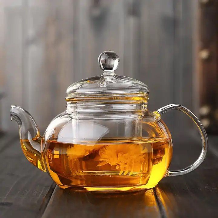 A transparent Crystal Clarity Teapot from My Store, made of high borosilicate glass and holding 600ml of amber-colored tea, rests on a wooden surface. The teapot includes a round handle, a curved spout, and a lid with a knob on top. Inside, a flowering tea bloom can be seen, adding an elegant touch to the brew.